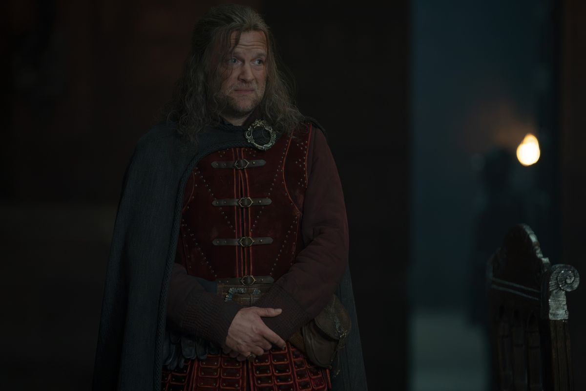 A man in a red studded doublet and dark cloak standing in a dim hall, hands clasped, looking off to one side
