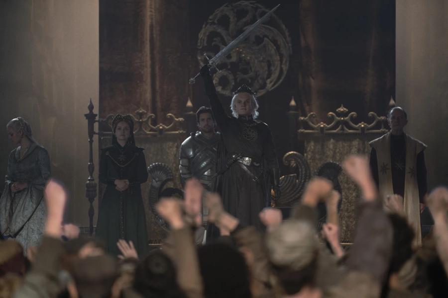 Tom Glynn-Carney as Aegon II, crowned, raising a sword above his head before a cheering crowd