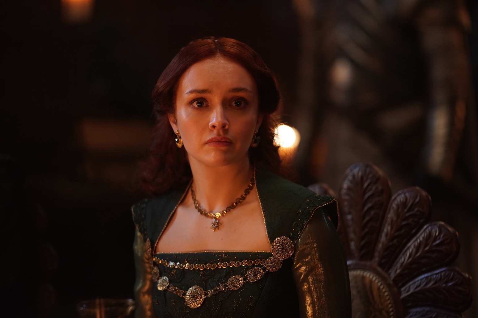 Alicent Hightower seated in a dark green gown, looking off to one side with a guarded expression