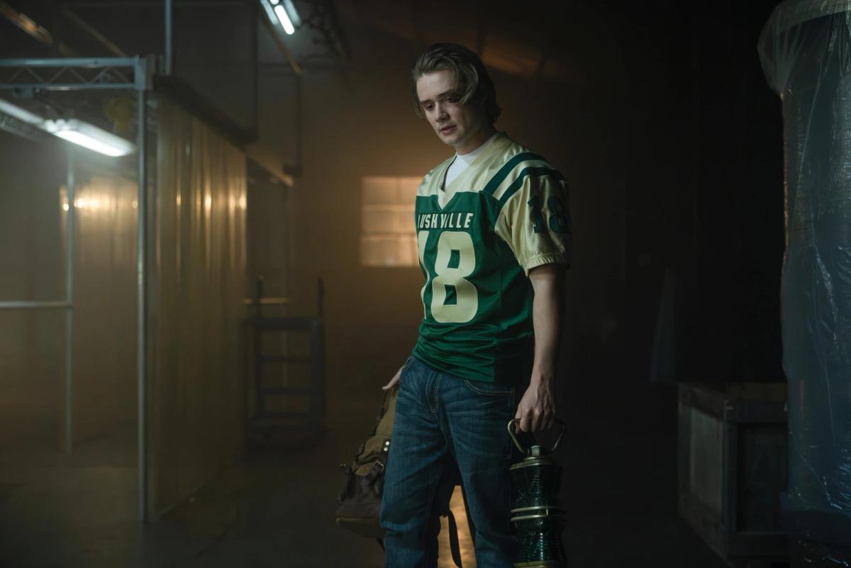 Shea Pritchard as Noah Macon in a Rushville number 18 football jersey, carrying a backpack and a green lantern along a dim, smoky corridor.