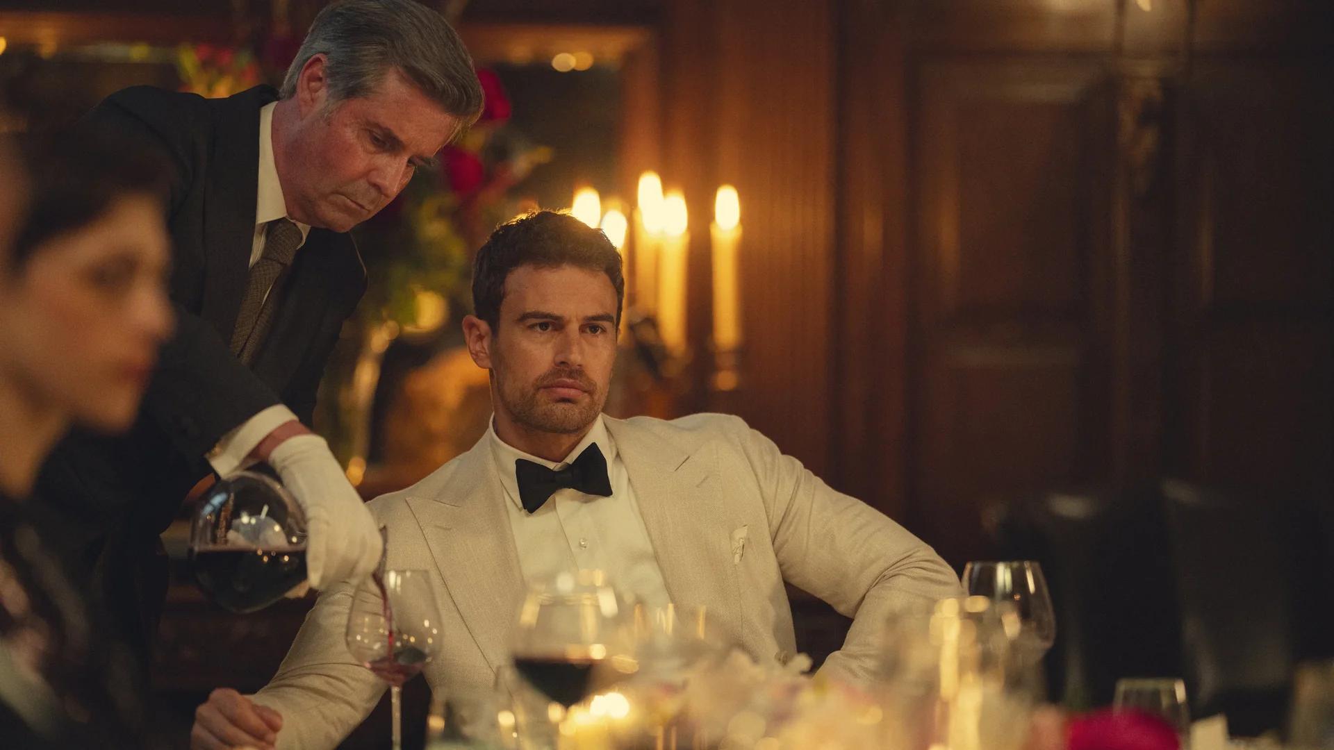 Theo James as Eddie Horniman in a white dinner jacket at a candlelit table while a butler pours wine over his shoulder.