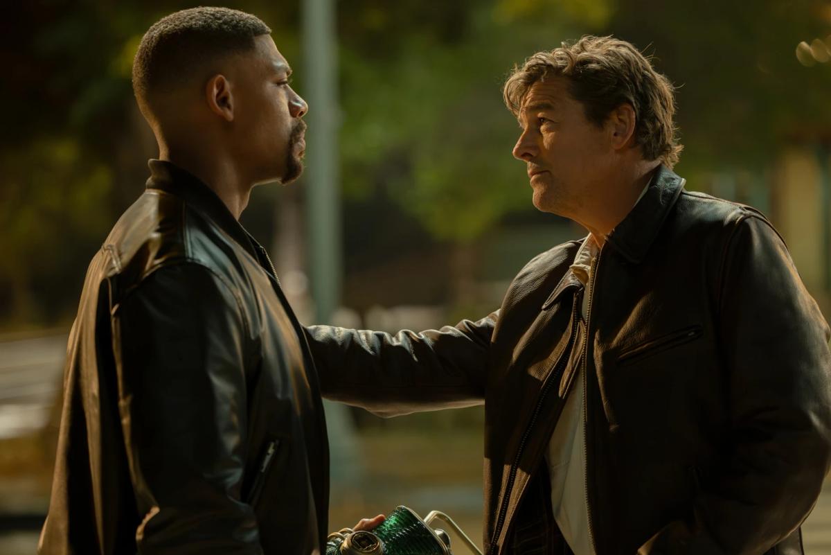 Kyle Chandler as the Halogram resting a hand on the shoulder of Aaron Pierre as John Stewart, who holds a green lantern, at night.