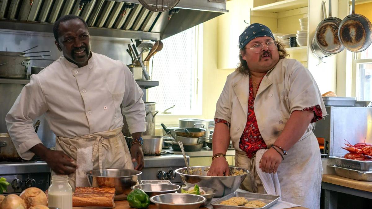 Michael Jibrin as Tyrone and Stavros Halkias as Stavros working a cluttered kitchen line, boiled lobsters stacked on a tray beside them.