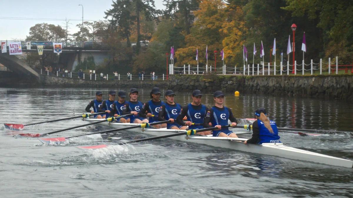 The Easton Prep eight racing past spectators on the bank, with Teagan Tao (Miku Martineau) facing her crew from the coxswain's seat.