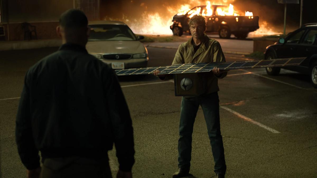 Kyle Chandler as Hal Jordan holding a retrieved satellite in a car park, a vehicle burning behind him.