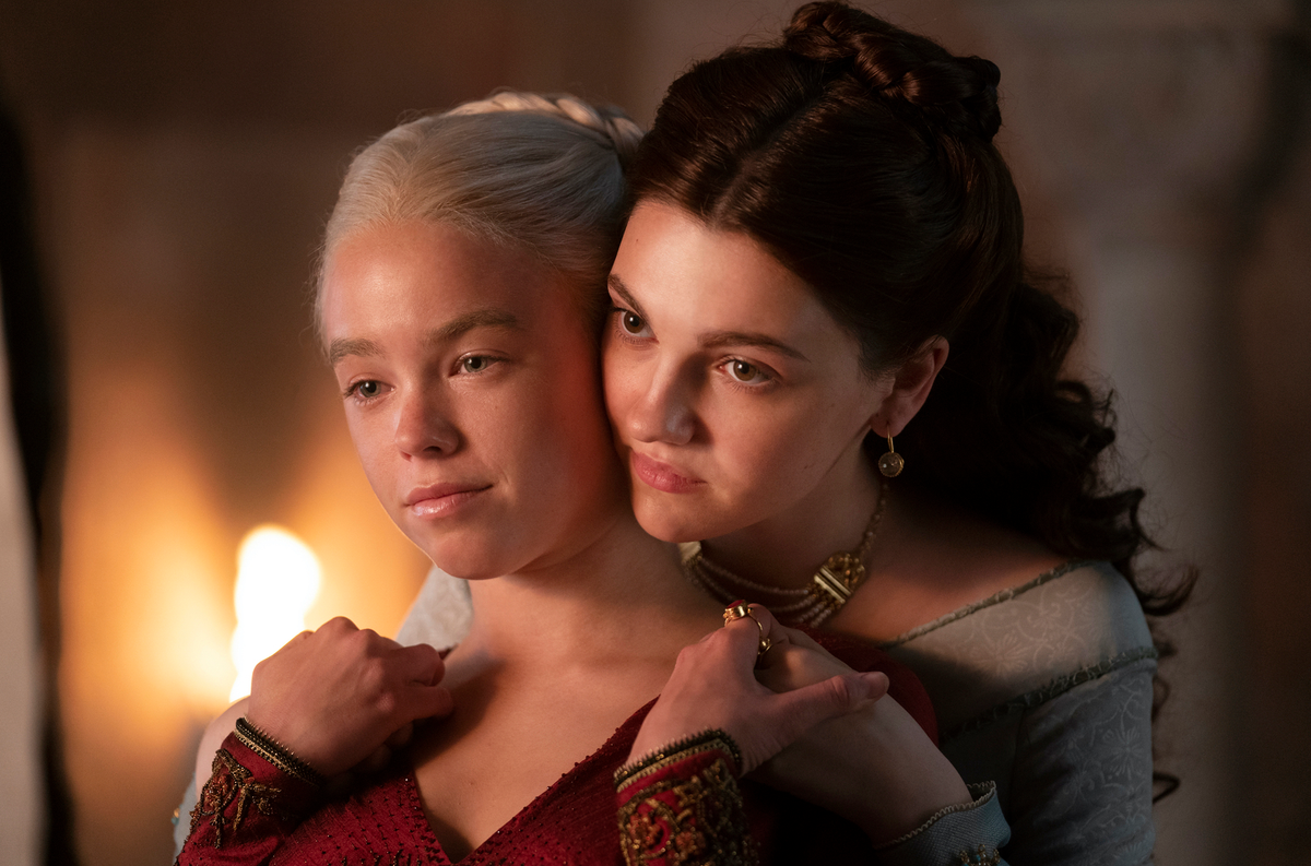 Young Rhaenyra and young Alicent standing close together by firelight, Alicent's arms around her shoulders