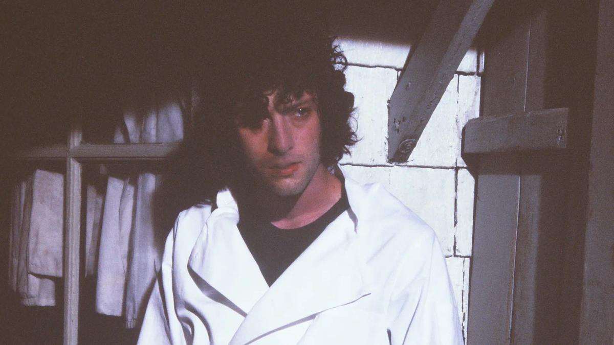 Dominic Sessa as Tony Bourdain pulling on an oversized chef jacket in a dim back room, looking off to one side.