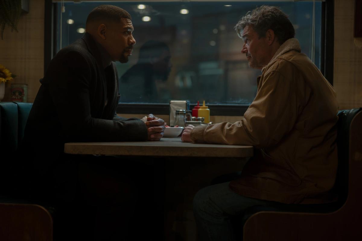 Aaron Pierre as John Stewart and Kyle Chandler as Hal Jordan sitting opposite each other in a café booth at night.