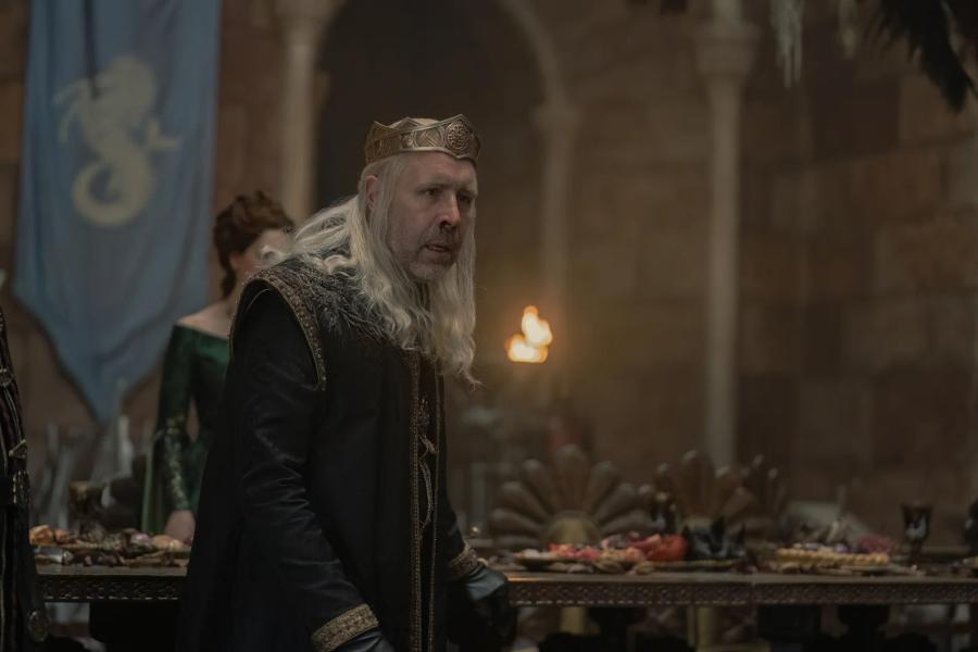 Paddy Considine as Viserys I, crowned, standing at a feast hall table