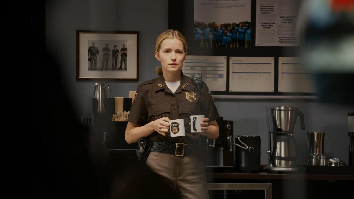 Willa Fitzgerald as Officer Roscoe Conklin carries two coffee mugs across the Margrave police station break room.