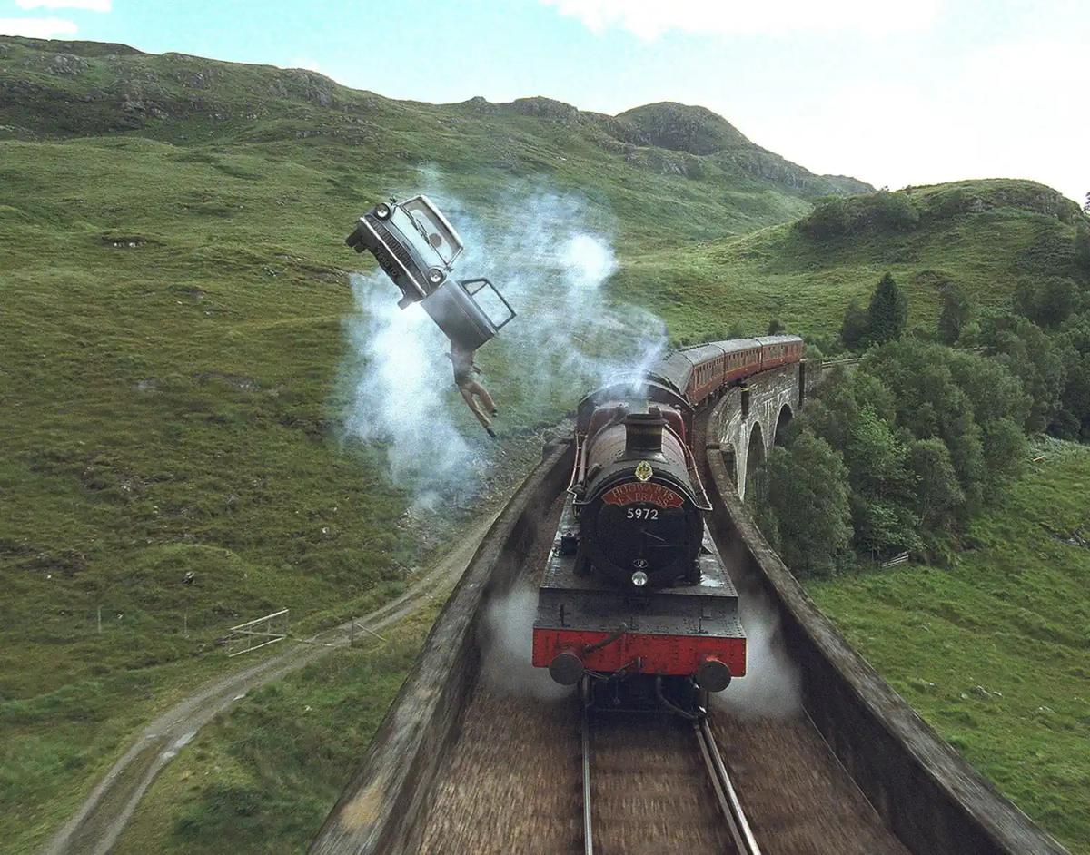 The flying Ford Anglia tips on its side above a viaduct beside the Hogwarts Express, with Harry Potter (Daniel Radcliffe) hanging from the open passenger door.