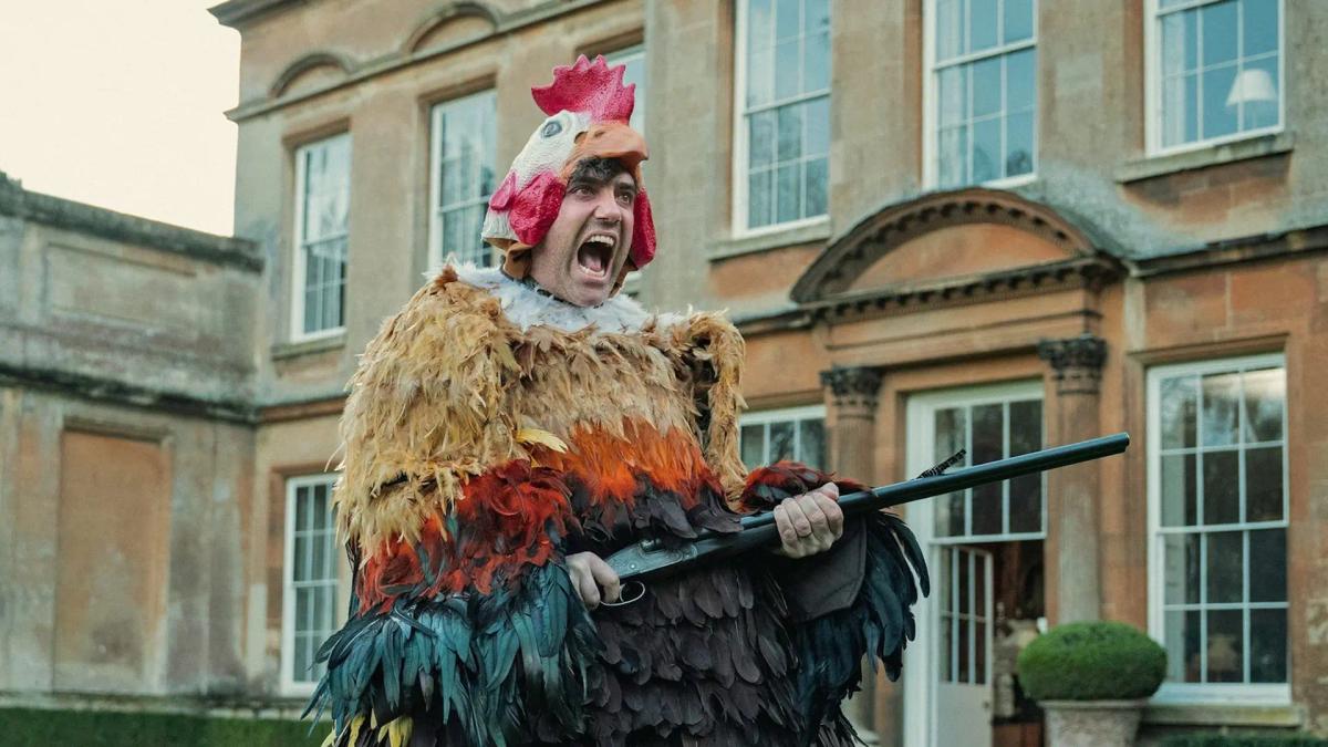 Daniel Ings as Freddy Horniman in a chicken costume outside Halstead Manor, mouth open mid-shout, holding a shotgun.
