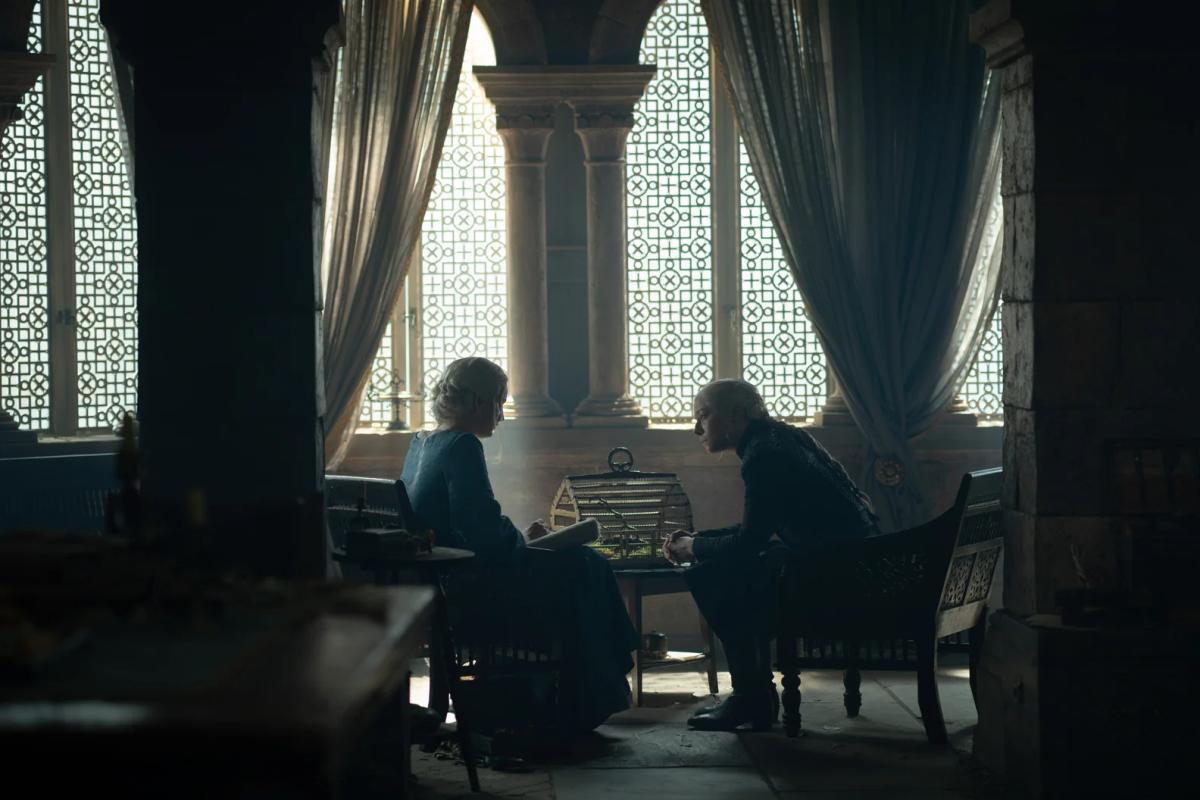 Phia Saban as Helaena Targaryen and Emma D’Arcy as Rhaenyra Targaryen sit facing each other by tall windows with a birdcage on the table between them