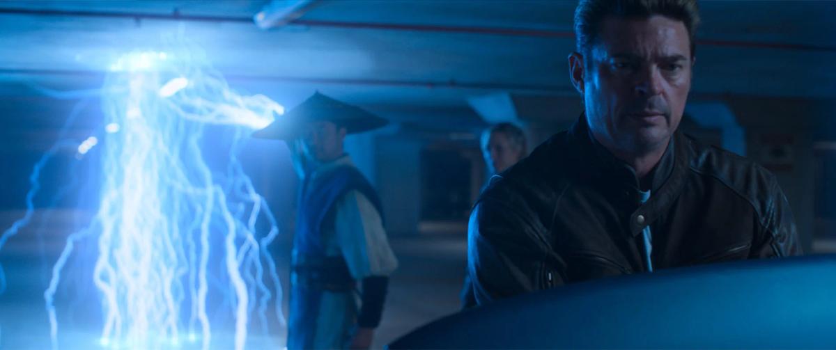Karl Urban as Johnny Cage in a dark car park, with Tadanobu Asano as Raiden in his wide hat and Jessica McNamee as Sonya Blade behind him, beside a column of blue lightning.