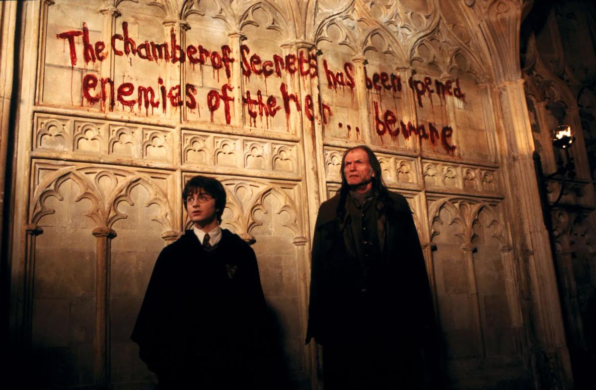 Harry Potter (Daniel Radcliffe) and Argus Filch (David Bradley) stand beneath a message daubed in red on the corridor wall reading that the Chamber of Secrets has been opened.