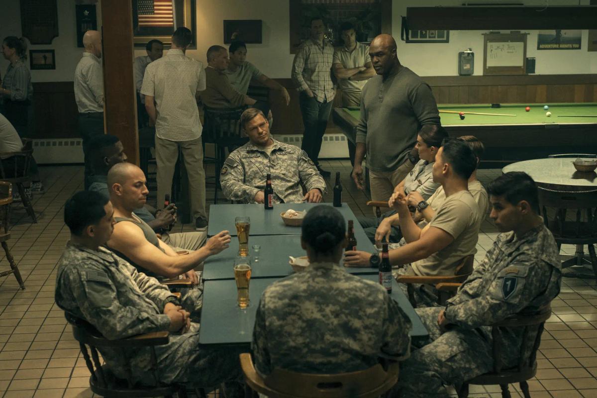 Flashback: Alan Ritchson as Jack Reacher sits in army fatigues at the head of a long table in a base bar, beers in front of him, the soldiers of the 110th Special Investigations Unit ranged around him and a pool table behind.