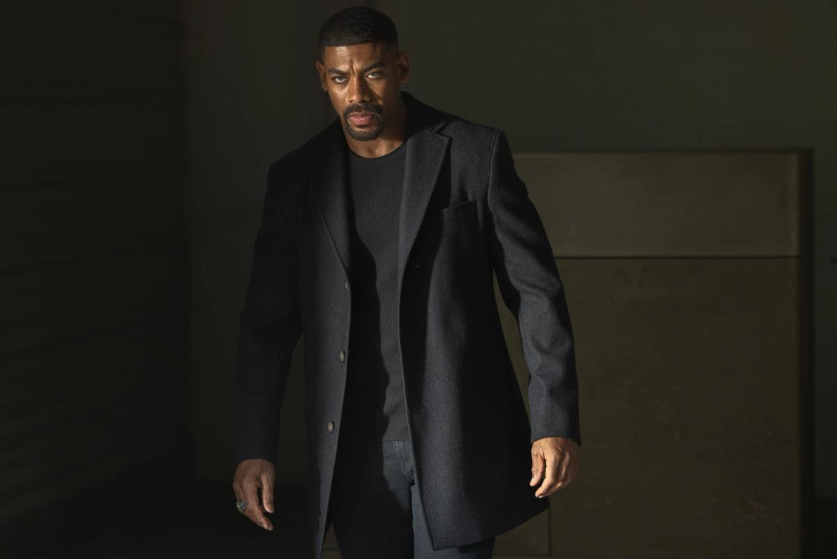 Aaron Pierre as John Stewart in a long dark overcoat, walking out of the shadows of a bare concrete room.