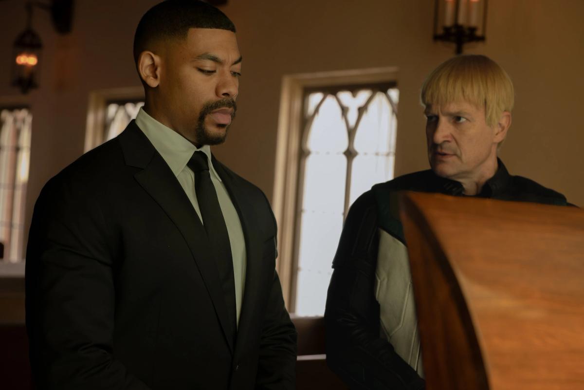 Aaron Pierre as John Stewart in a black suit and Nathan Fillion as Guy Gardner in his Green Lantern uniform, standing beside a coffin.