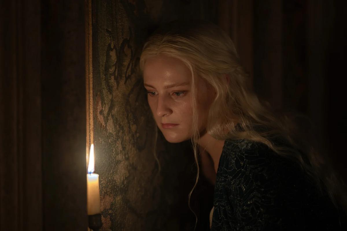 Phia Saban as Helaena Targaryen lit by a single candle, looking down, beside a painted wall