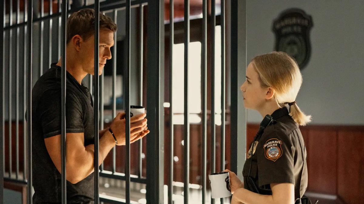 Alan Ritchson as Jack Reacher stands behind the bars of the Margrave police holding cell, passing a coffee mug through to Willa Fitzgerald as Officer Roscoe Conklin.