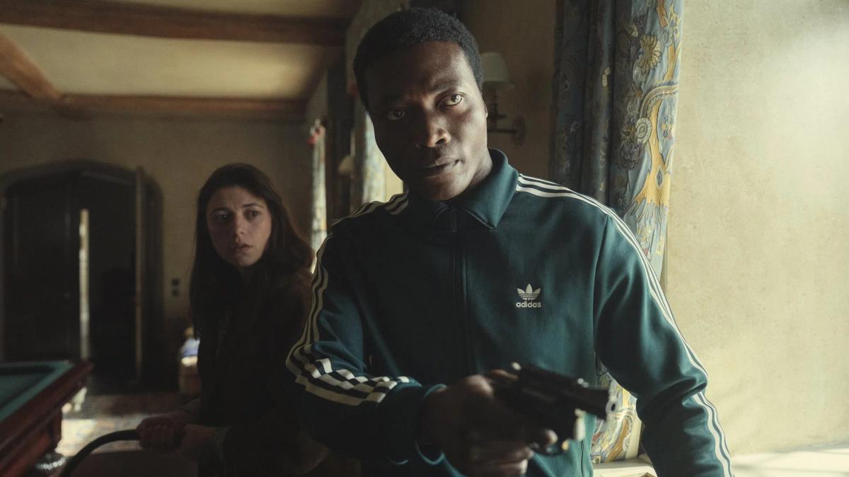 Benjamin Clementine as Nanny levelling a revolver inside Halstead Manor, with Jasmine Blackborow as Charly Horniman behind him.
