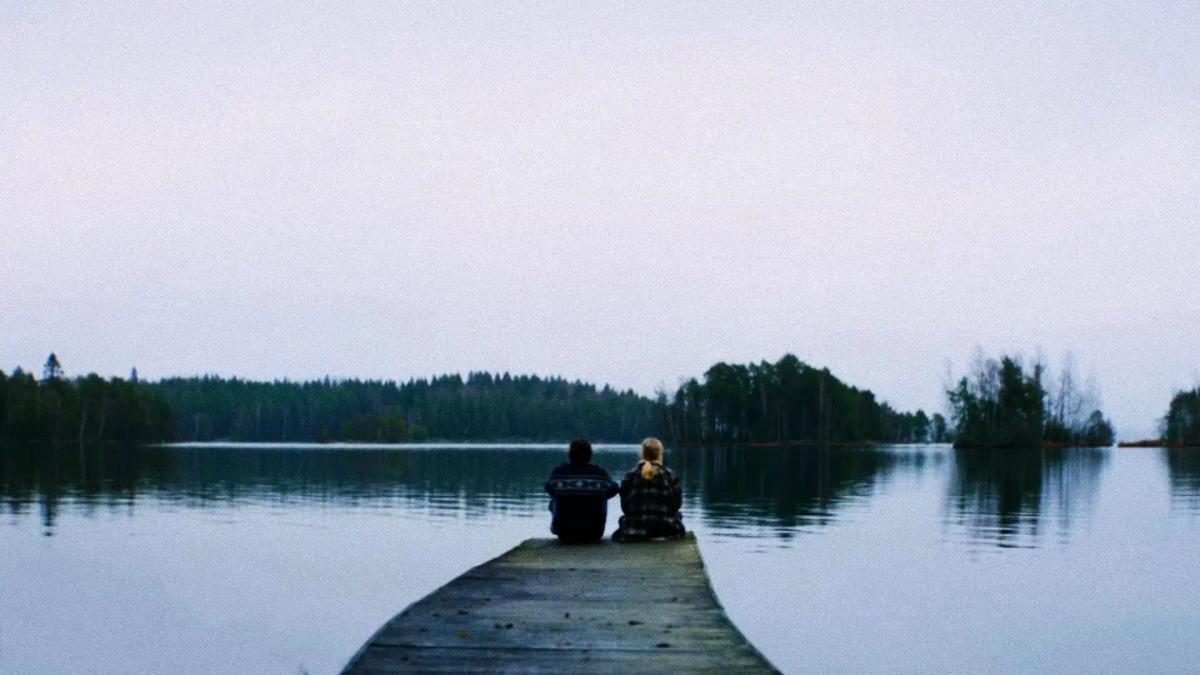 Jason Segel as Dan and Samara Weaving as Lisa sitting apart at the end of a jetty on a still lake at dusk.