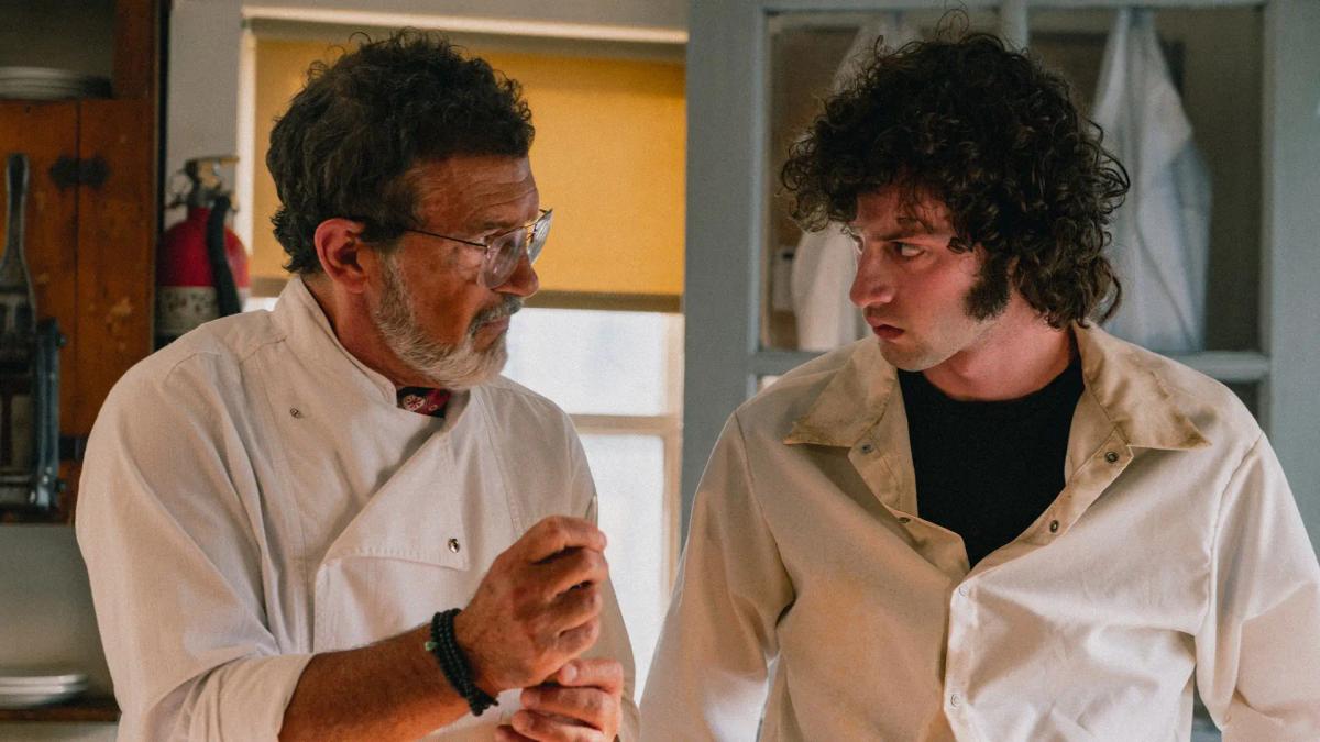 Antonio Banderas as Chef, in whites and wire-rimmed glasses, turning to look at Dominic Sessa as Tony Bourdain across the kitchen.