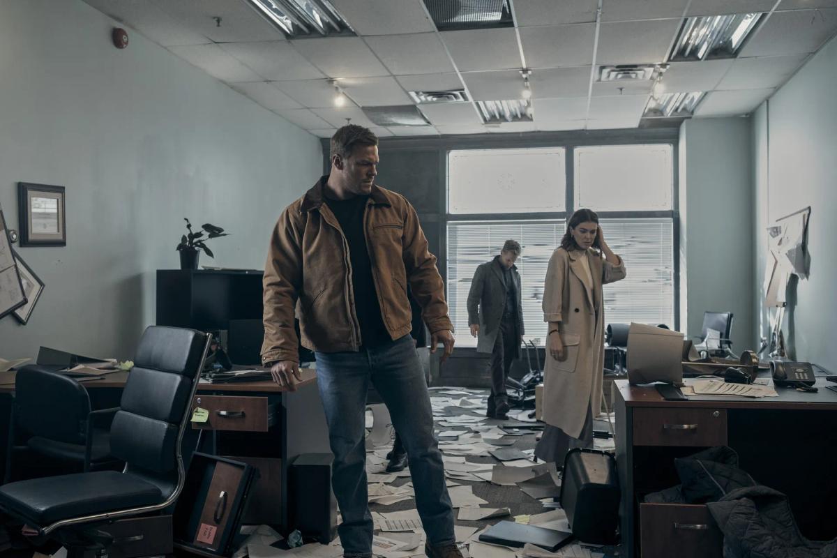 Alan Ritchson as Jack Reacher stands in an office strewn with paper and overturned chairs, with Shaun Sipos as David O'Donnell and Serinda Swan as Karla Dixon searching the room behind him.
