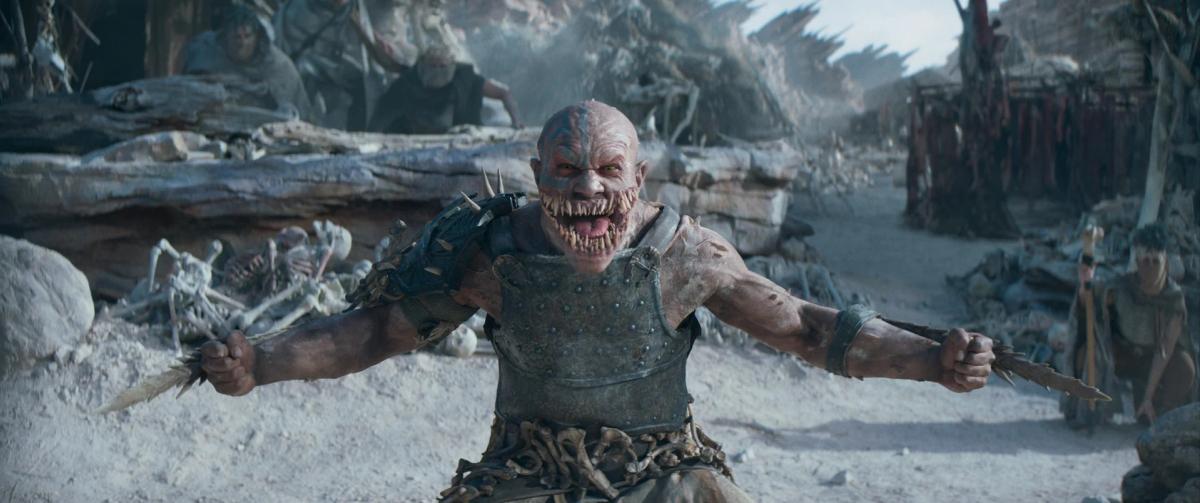 CJ Bloomfield as Baraka, blades out from both arms, snarling in a pale, bone-strewn village.