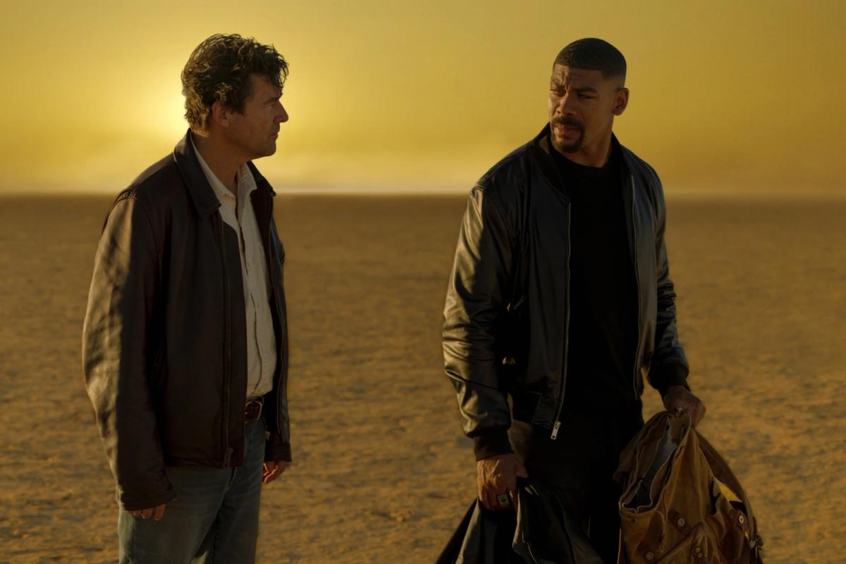 Kyle Chandler as the Halogram and Aaron Pierre as John Stewart, holding a backpack, on a flat yellow desert under a hazy yellow sky.