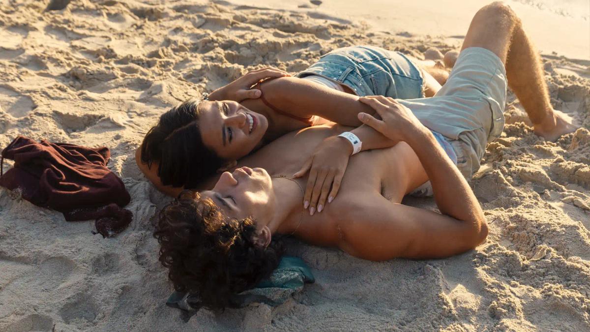 Maia Reficco as Oriah "Ry" Pera and Fernando Lindez as Julián García lying together on the sand at sunset.