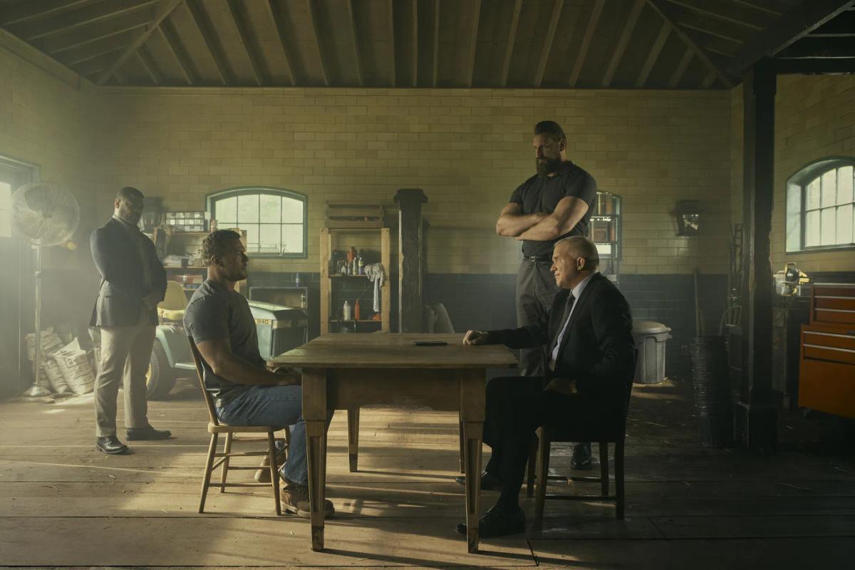 Alan Ritchson as Jack Reacher sits across a wooden table from Anthony Michael Hall as Zachary Beck in a brick garage, while Olivier Richters as Paulie stands behind Zachary Beck with his arms folded and another man watches from the side.