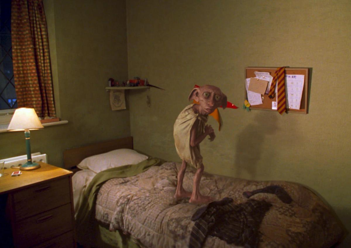 Dobby (voiced by Toby Jones) stands on the bed in Harry Potter's small bedroom at Privet Drive, wearing a pillowcase.