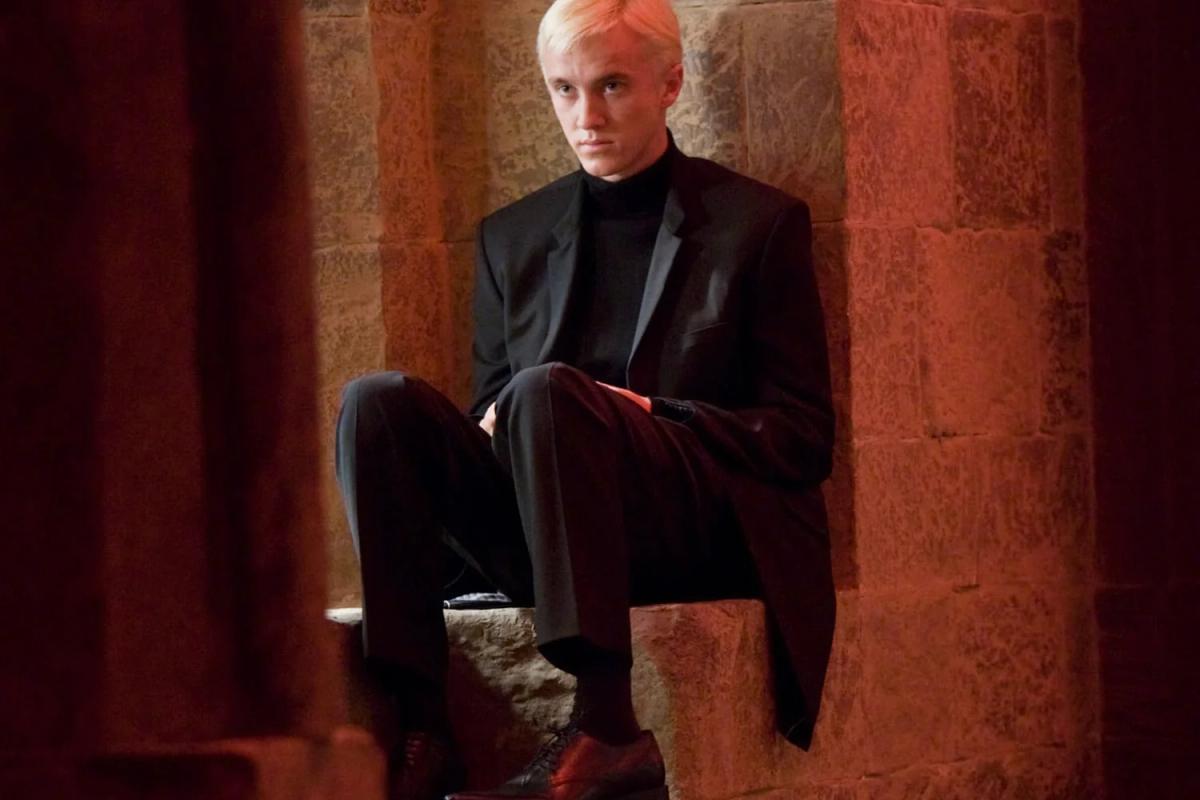 Tom Felton as Draco Malfoy sitting alone on a window ledge in a red corridor