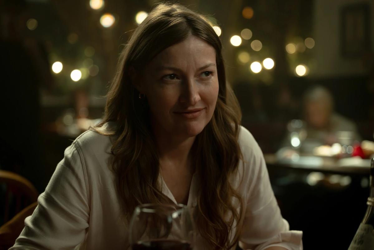 Kelly Macdonald as Kerry Kane, smiling at a restaurant table with a glass of red wine in front of her.