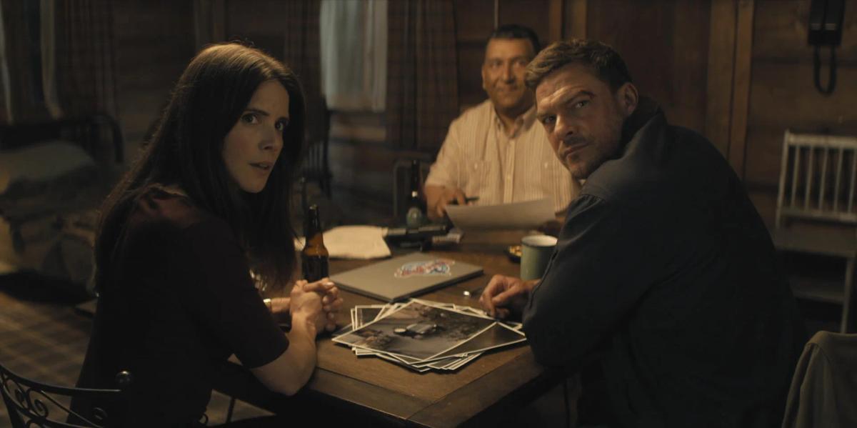Sonya Cassidy as Susan Duffy and Alan Ritchson as Jack Reacher turn to look over their shoulders from a table spread with photographs, a laptop and beer bottles, with Roberto Montesinos as Guillermo Villanueva sitting behind them holding a sheet of paper.