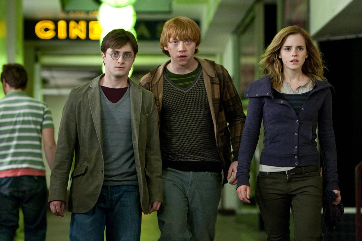 Harry Potter (Daniel Radcliffe), Ron Weasley (Rupert Grint) and Hermione Granger (Emma Watson) walk down a lit Muggle street past a cinema, still in the clothes they escaped in.