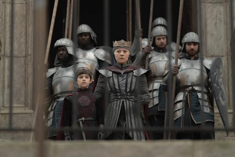 Emma D’Arcy as Rhaenyra Targaryen, crowned and in mail, holding the hand of a young boy in Targaryen colours, with guards behind them