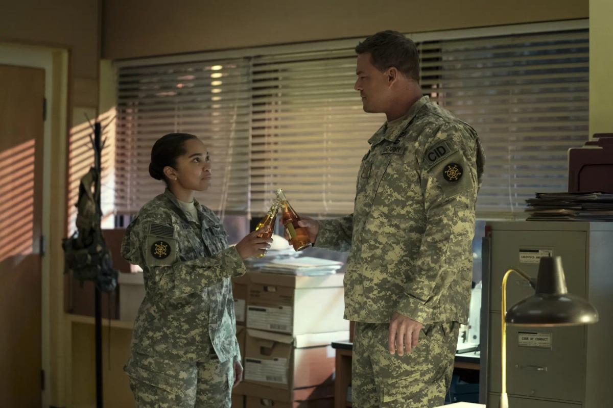 Flashback: Mariah Robinson as Dominique Kohl and Alan Ritchson as Jack Reacher, both in Army camouflage uniforms, clink bottles in an office with the blinds drawn.