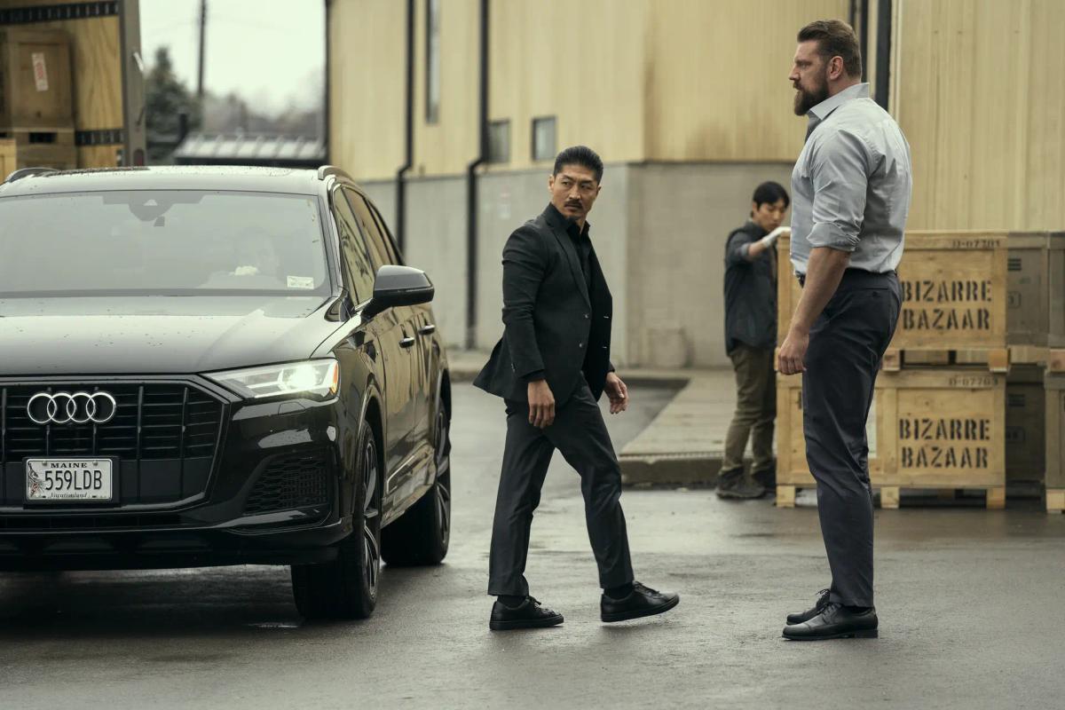 Brian Tee as Francis Xavier Quinn, in a black suit, crosses a wet loading yard from a black SUV toward Olivier Richters as Paulie, who stands beside wooden crates stencilled Bizarre Bazaar.