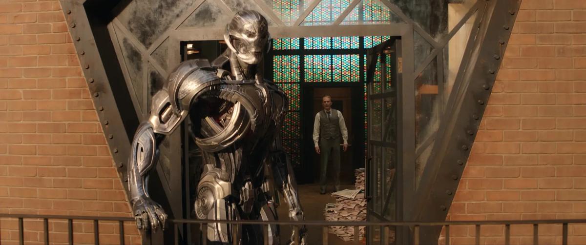 Ultron in his metal robot body leans on a balcony rail while Paul Bettany as Vision watches from a doorway behind him in VisionQuest