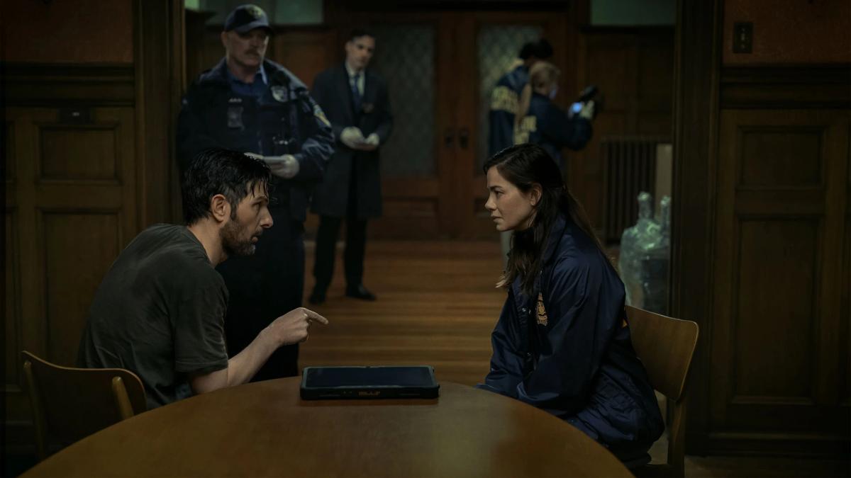 Adam Scott as Tom Kennedy sitting across a table from Michelle Monaghan as Amanda Beck, with officers standing behind them.