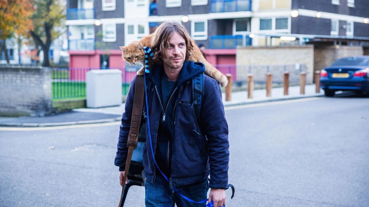 Luke Treadaway as James Bowen walking a London street with Bob the cat draped across his shoulders and a guitar case in his hand.