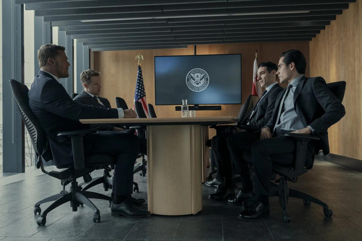 Alan Ritchson as Jack Reacher and Shaun Sipos as David O'Donnell sit in suits at a conference table facing two officials, beneath a screen carrying the U.S. Department of Homeland Security seal and beside a United States flag.