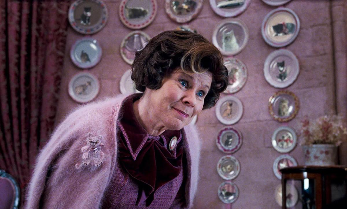 Dolores Umbridge (Imelda Staunton) in a pink cardigan, smiling in front of a wall of decorative kitten plates.