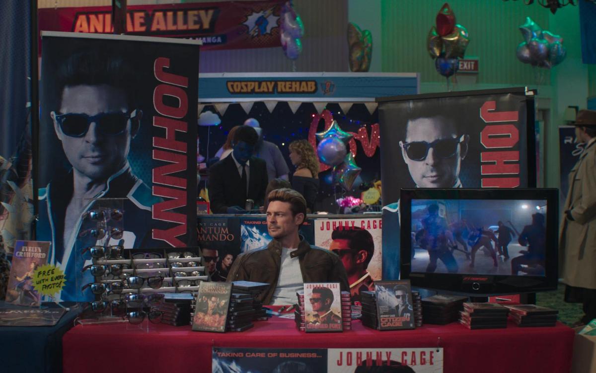 Karl Urban as Johnny Cage sitting at a fan convention table covered in his old films, under posters of himself in sunglasses.
