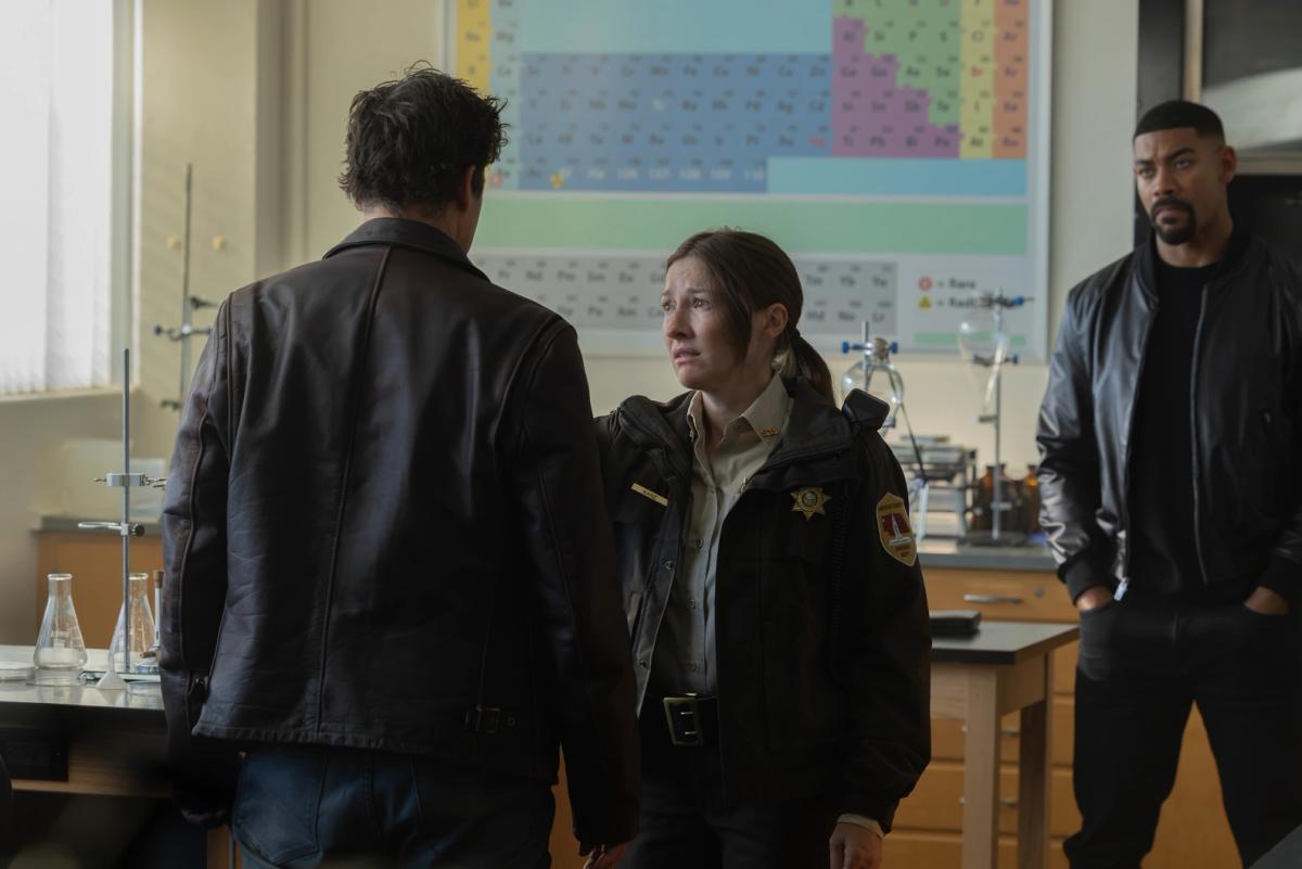 Kelly Macdonald as Kerry Kane, in uniform and close to tears, facing Kyle Chandler's Halogram, seen from behind, in a school science classroom while Aaron Pierre as John Stewart watches.