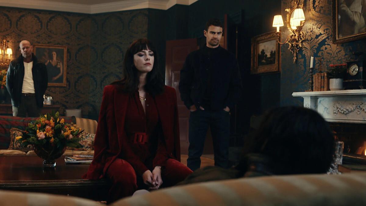 Kaya Scodelario as Susie Glass in a red suit seated in the Halstead drawing room, with Theo James as Eddie Horniman standing behind her.