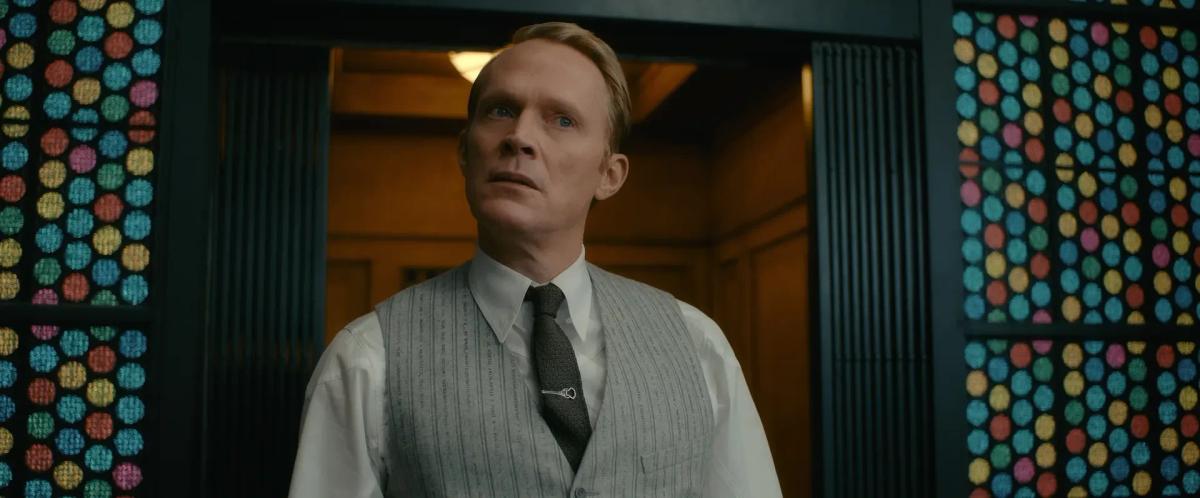 Paul Bettany as Vision in a waistcoat and tie, standing in front of an elevator framed by coloured glass panels in VisionQuest
