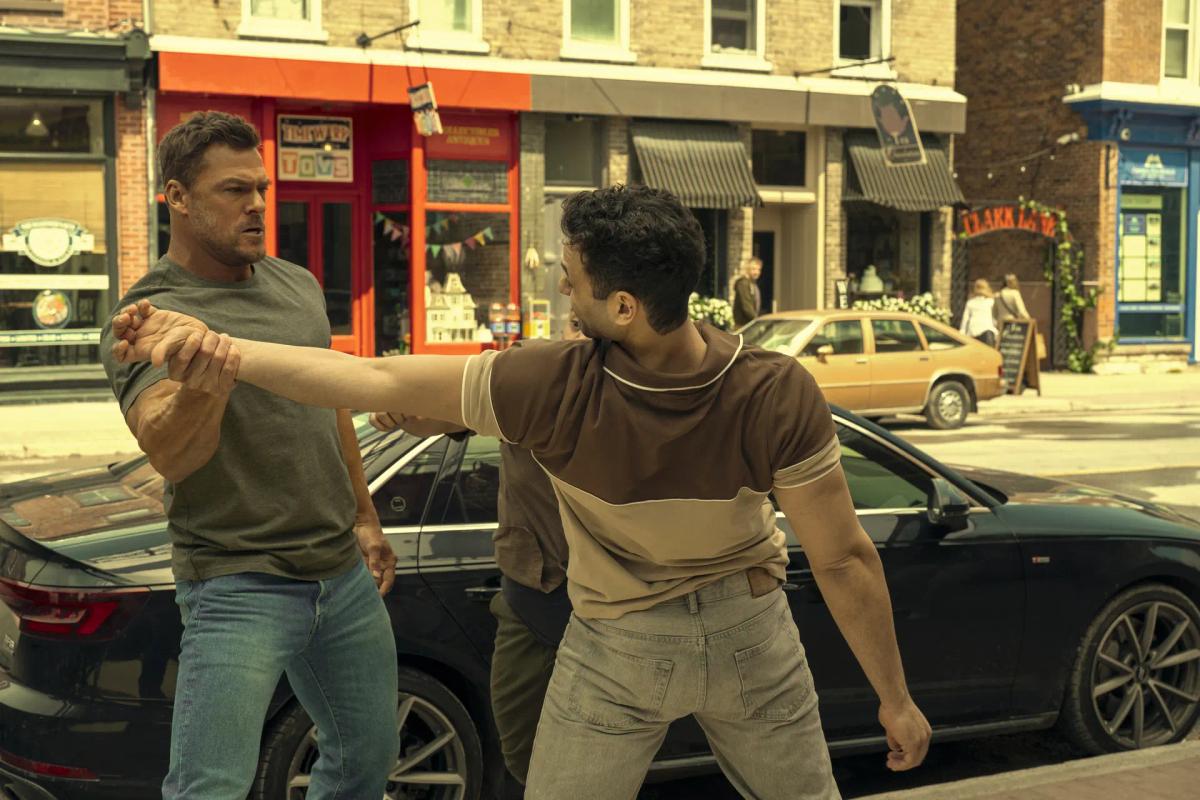 Alan Ritchson as Jack Reacher grabs the outstretched arm of a young man in a brown polo shirt on a small-town street, with a second man close behind them and a black car parked alongside.